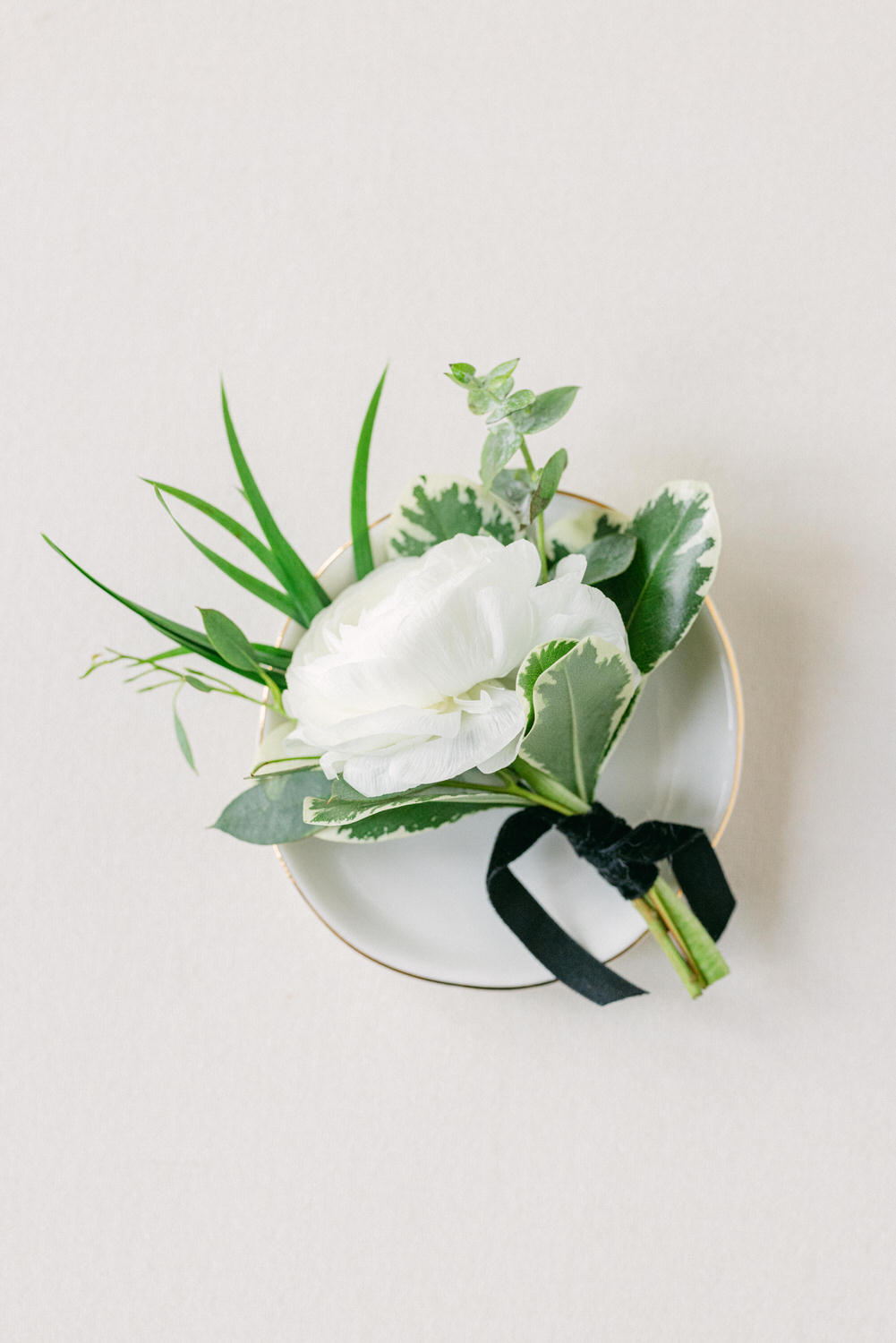 A beautiful white flower with greenery arranged on a delicate plate, tied with a black ribbon against a light background.