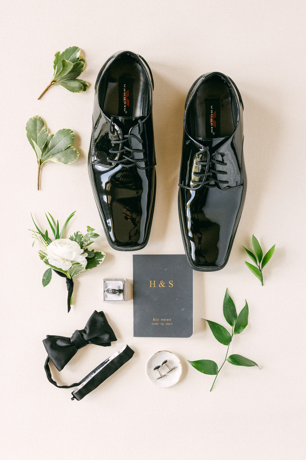 A flat lay of polished black dress shoes, a bow tie, rings, a vow notebook, a boutonnière, and greenery, arranged on a soft beige background.