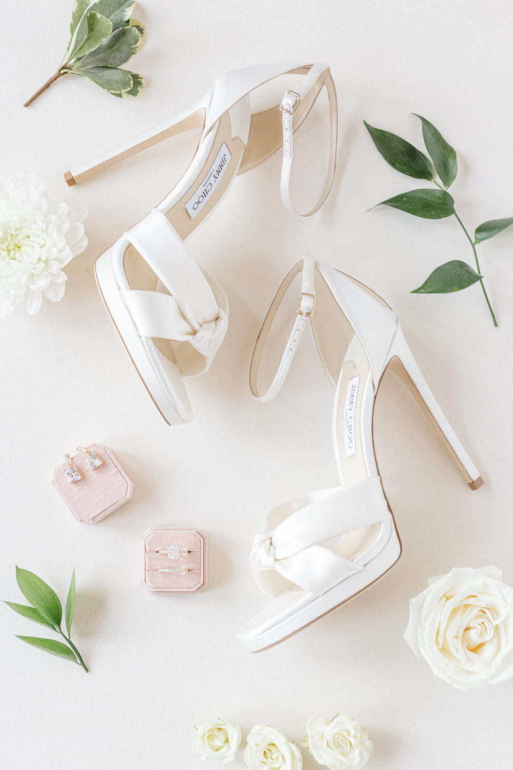 A pair of stylish white high-heeled sandals paired with delicate jewelry boxes and fresh white flowers, arranged on a soft background.