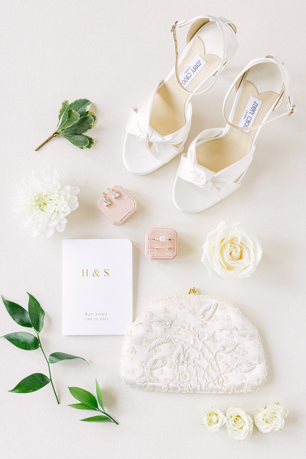 A flat lay of wedding essentials, including white satin heels, floral accents, jewelry boxes, a vow booklet, and a decorative clutch, arranged on a soft background.