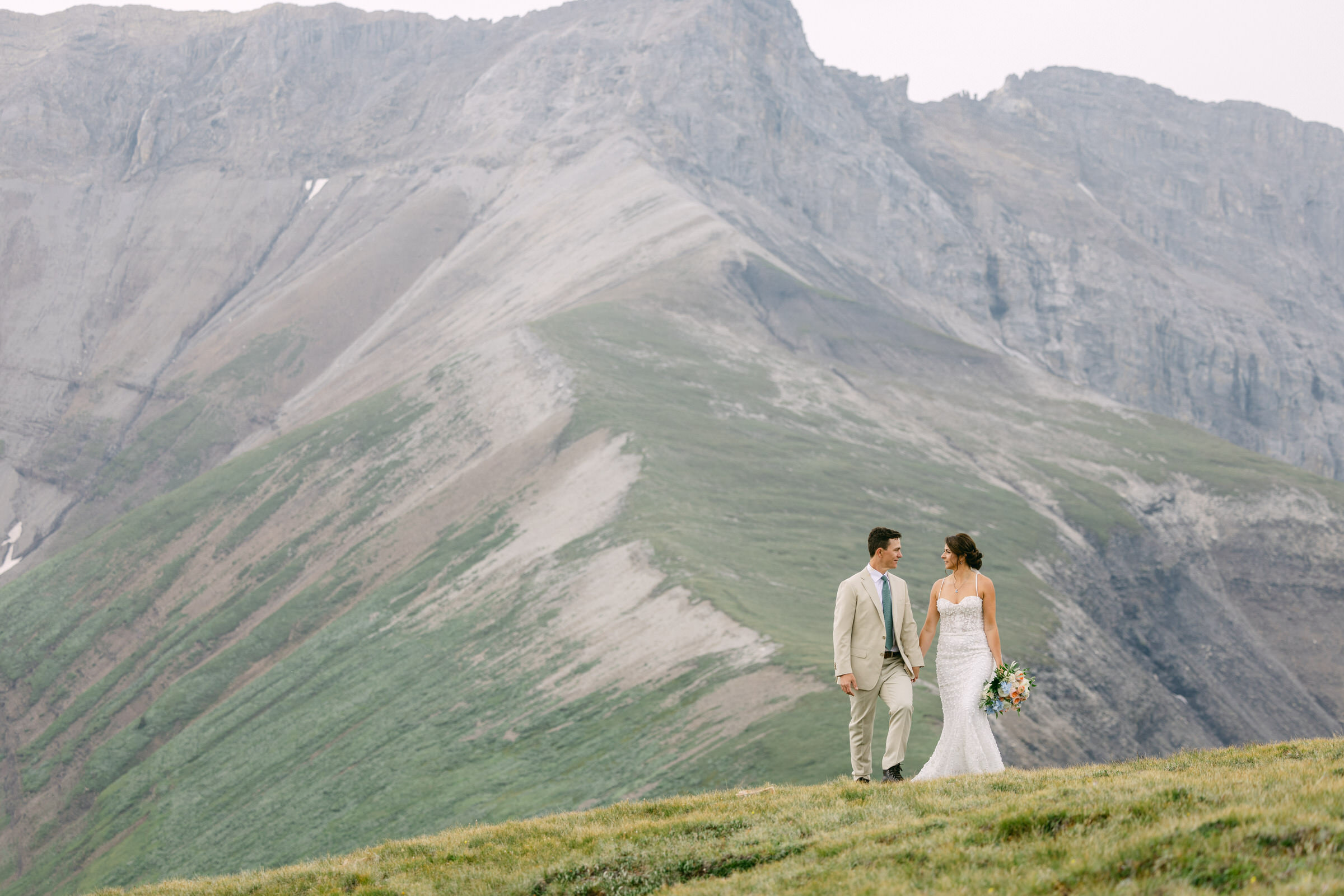 A couple walks hand in hand on a grassy hillside, with majestic mountains in the background, during their wedding ceremony.