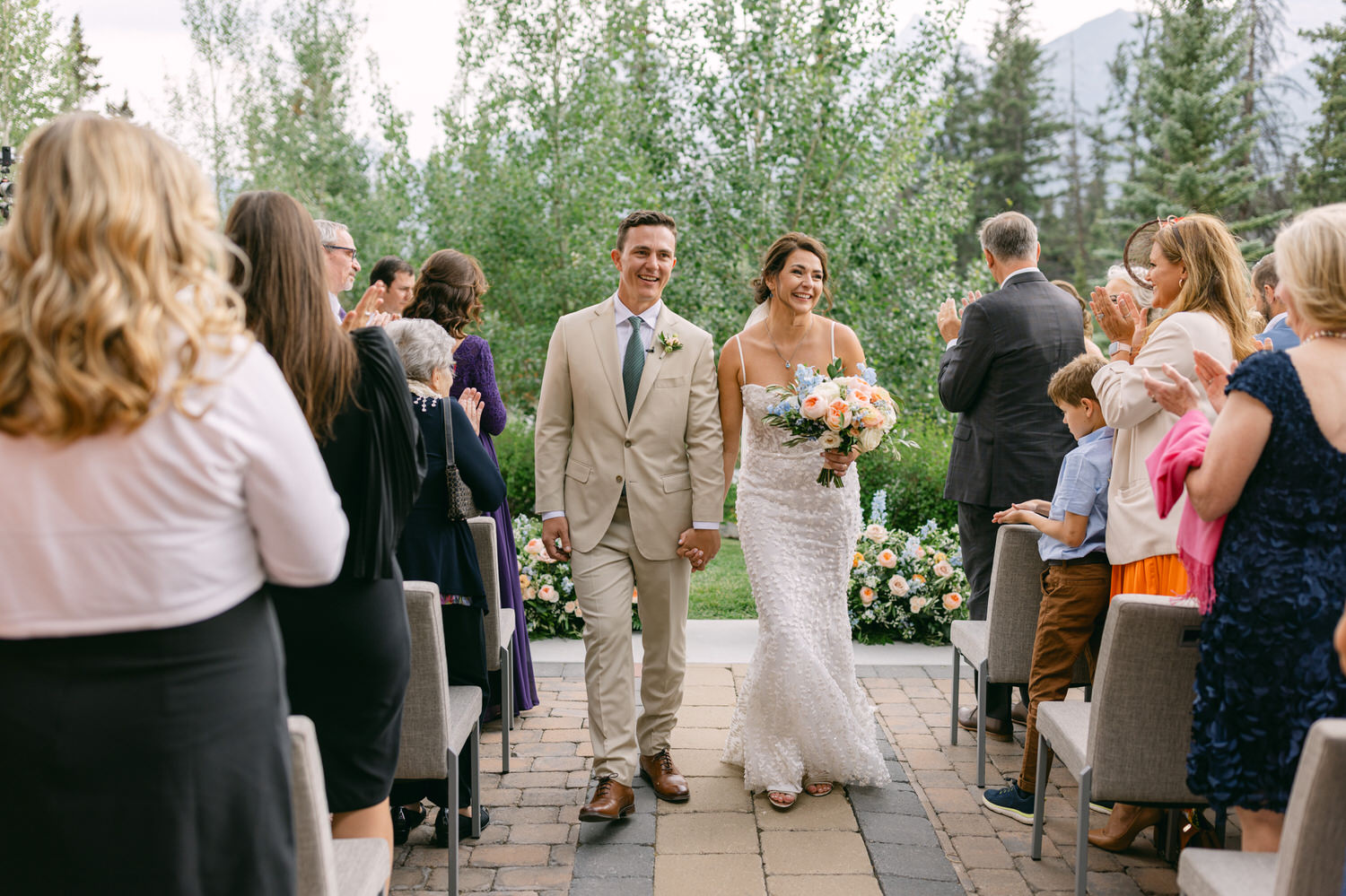 A newlywed couple walks hand in hand down the aisle, smiling and surrounded by applauding guests in a picturesque outdoor setting.