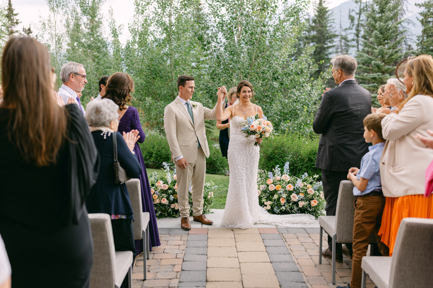 A joyful couple celebrating their marriage, surrounded by applauding guests and a beautiful outdoor setting with lush greenery and floral decorations.