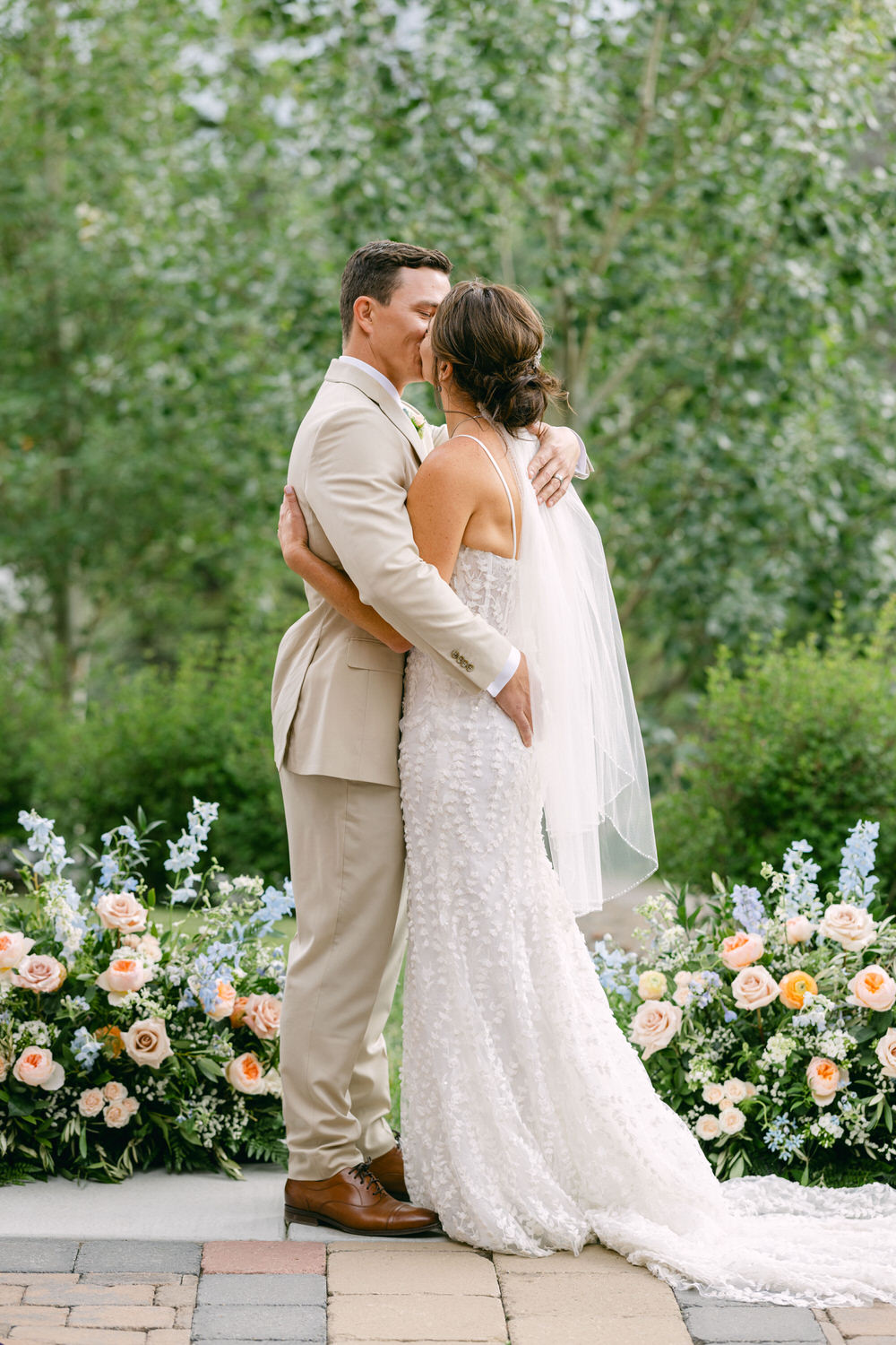 A couple shares a tender moment, kissing in front of a lush green backdrop adorned with floral arrangements.