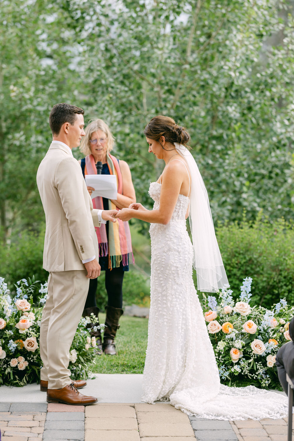 A bride and groom exchange vows during an outdoor wedding ceremony, with a celebrant reading in the background and floral arrangements nearby.