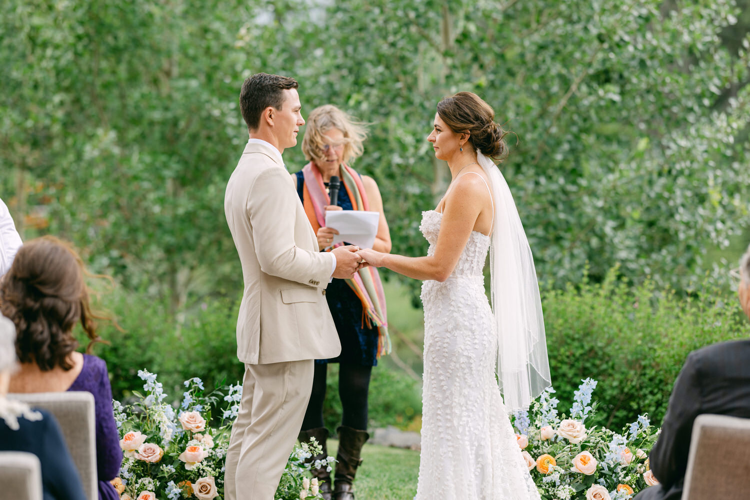 A bride and groom exchange vows during their outdoor wedding ceremony, surrounded by lush greenery and floral arrangements.