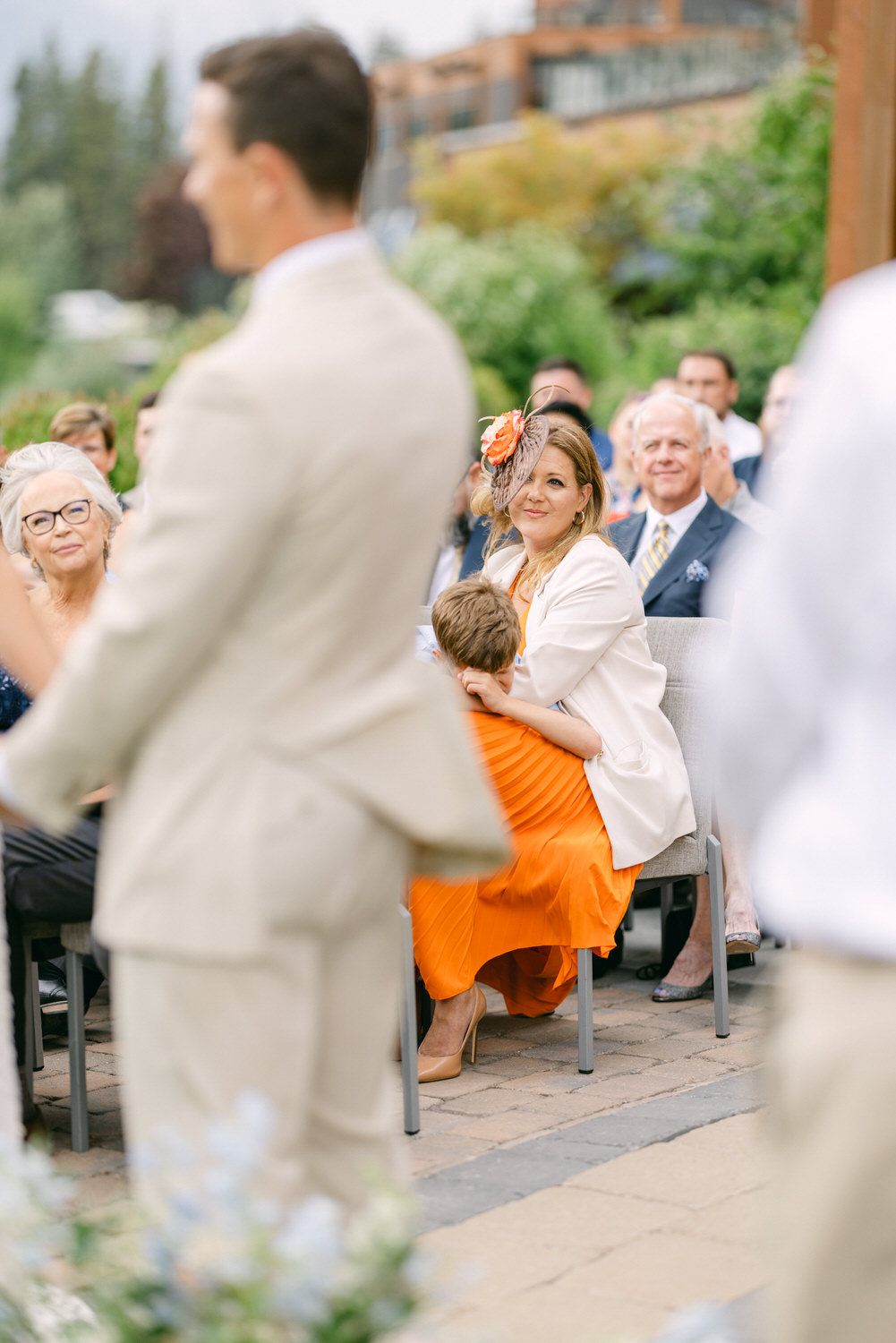 A woman in an orange dress smiles while holding a child, surrounded by seated guests in a festive outdoor setting.