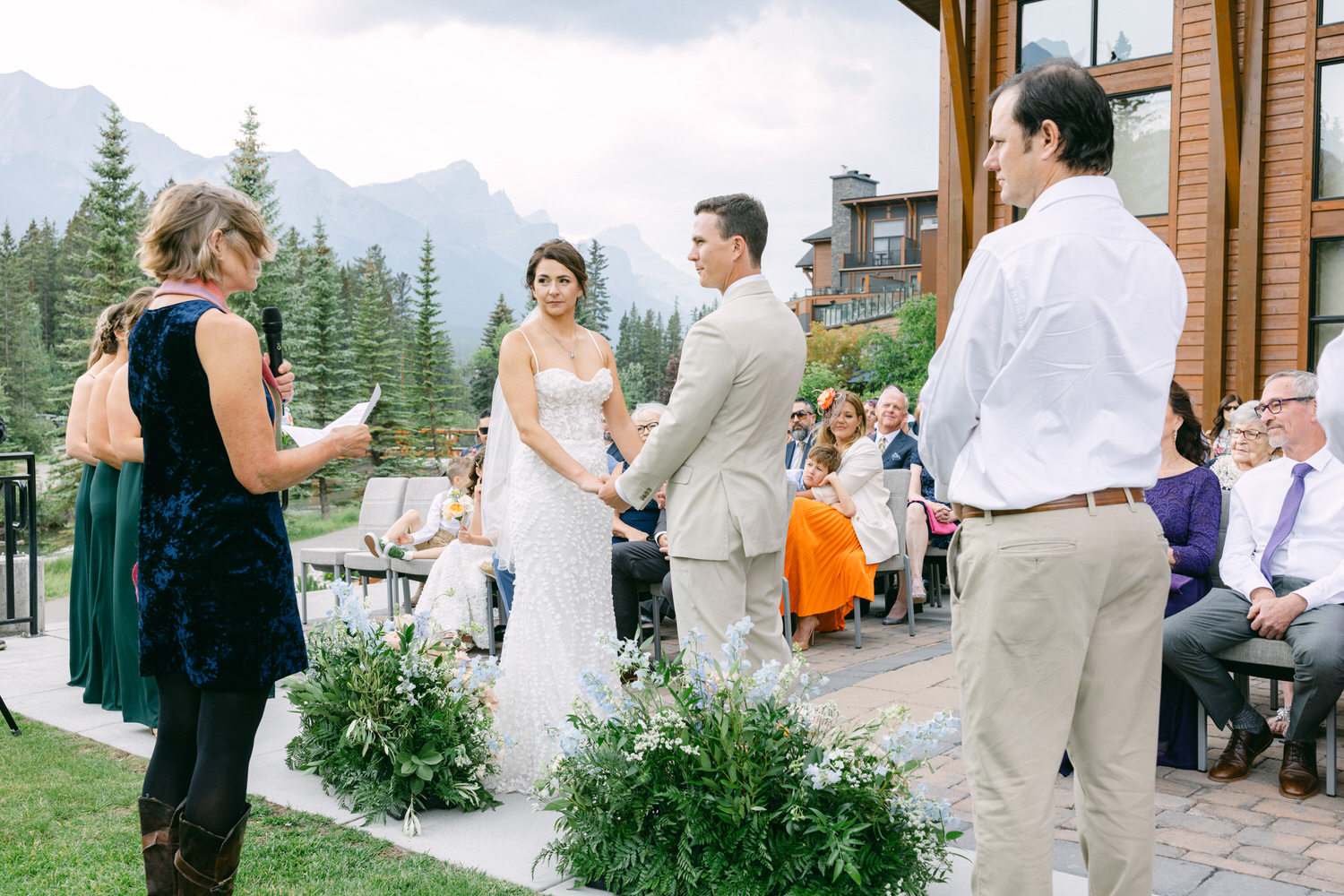 A bride and groom exchanging vows during an outdoor wedding ceremony, surrounded by guests and a scenic mountain view.