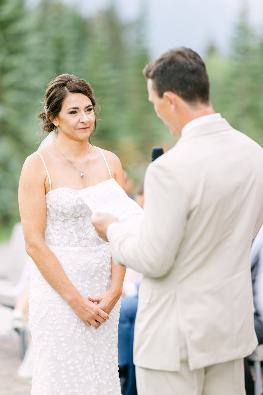 A bride listens attentively as her partner reads vows during an outdoor wedding ceremony surrounded by nature.