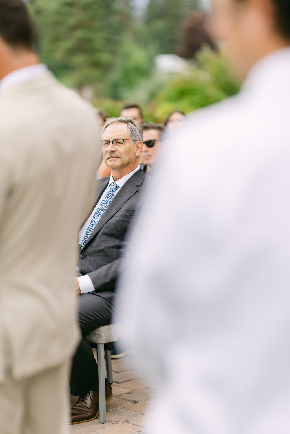 A well-dressed man sitting among guests at an outdoor event, intently watching the ceremony with a backdrop of greenery and blurred figures.