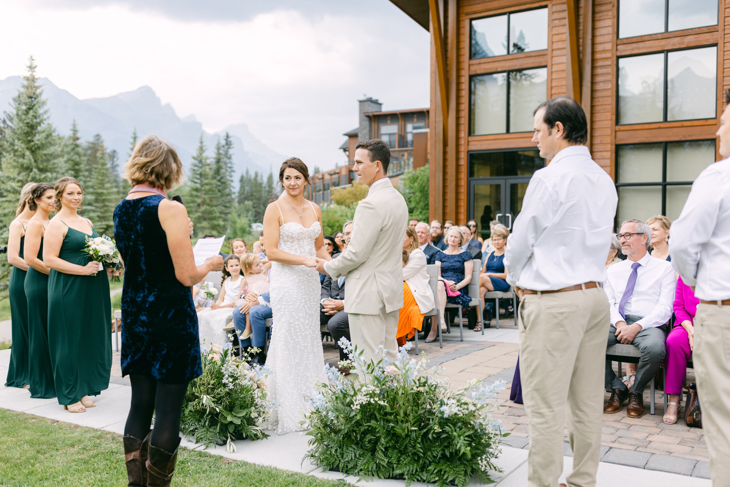 A bride and groom exchange vows during an outdoor wedding ceremony, surrounded by guests and nature.