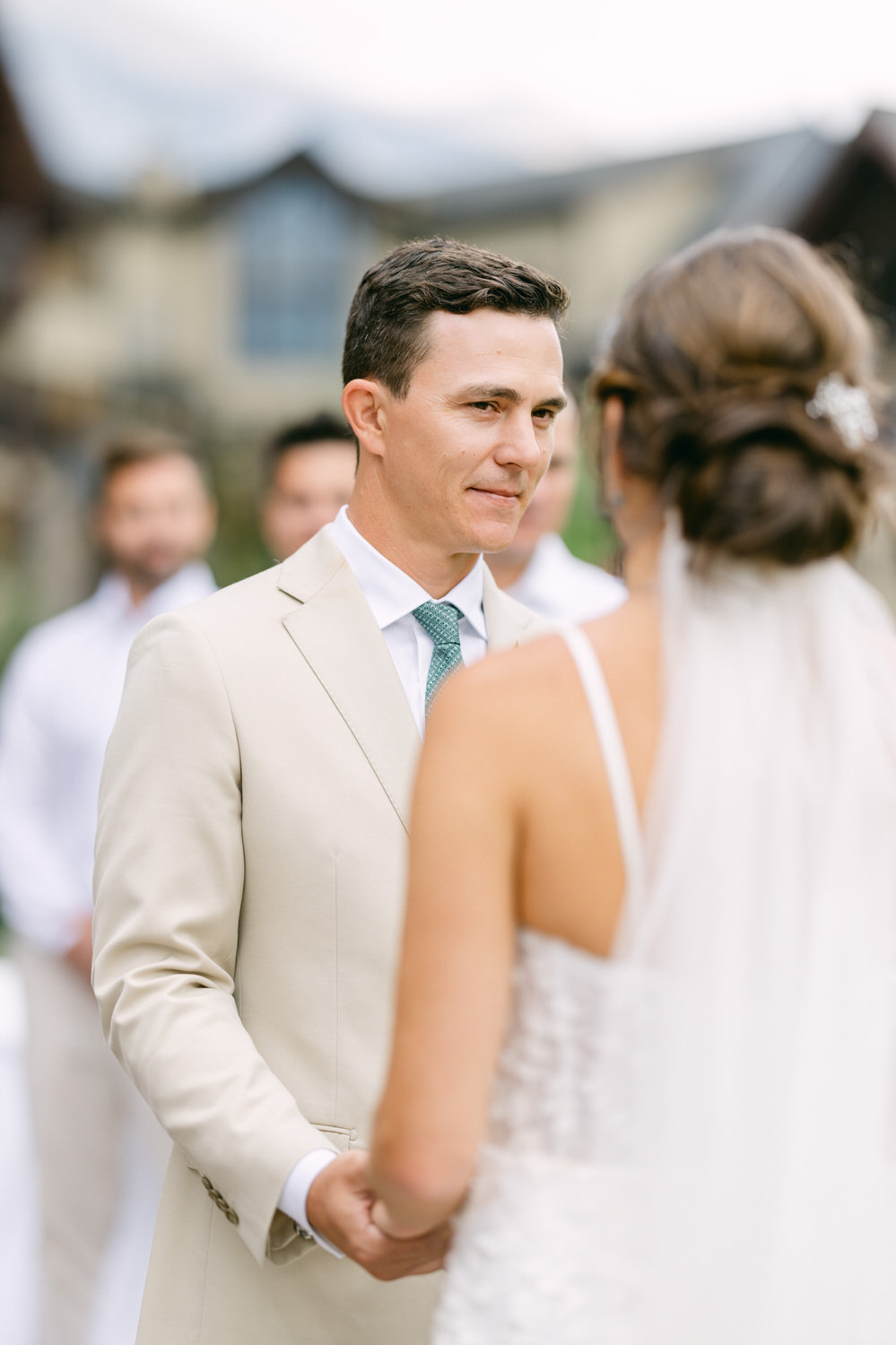 Wedding Ceremony Moment::A groom in a beige suit gazes intently at his bride during their wedding ceremony, surrounded by a serene outdoor setting.