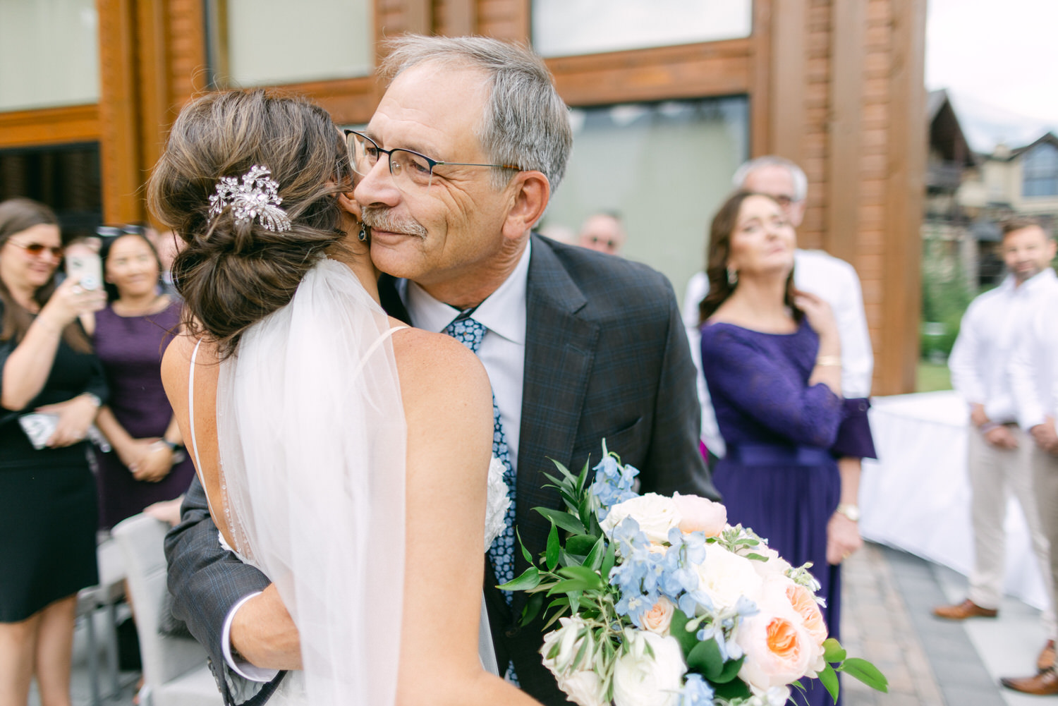 A joyful moment between a bride and her father, sharing an emotional hug during a wedding ceremony, with guests capturing the memory in the background.