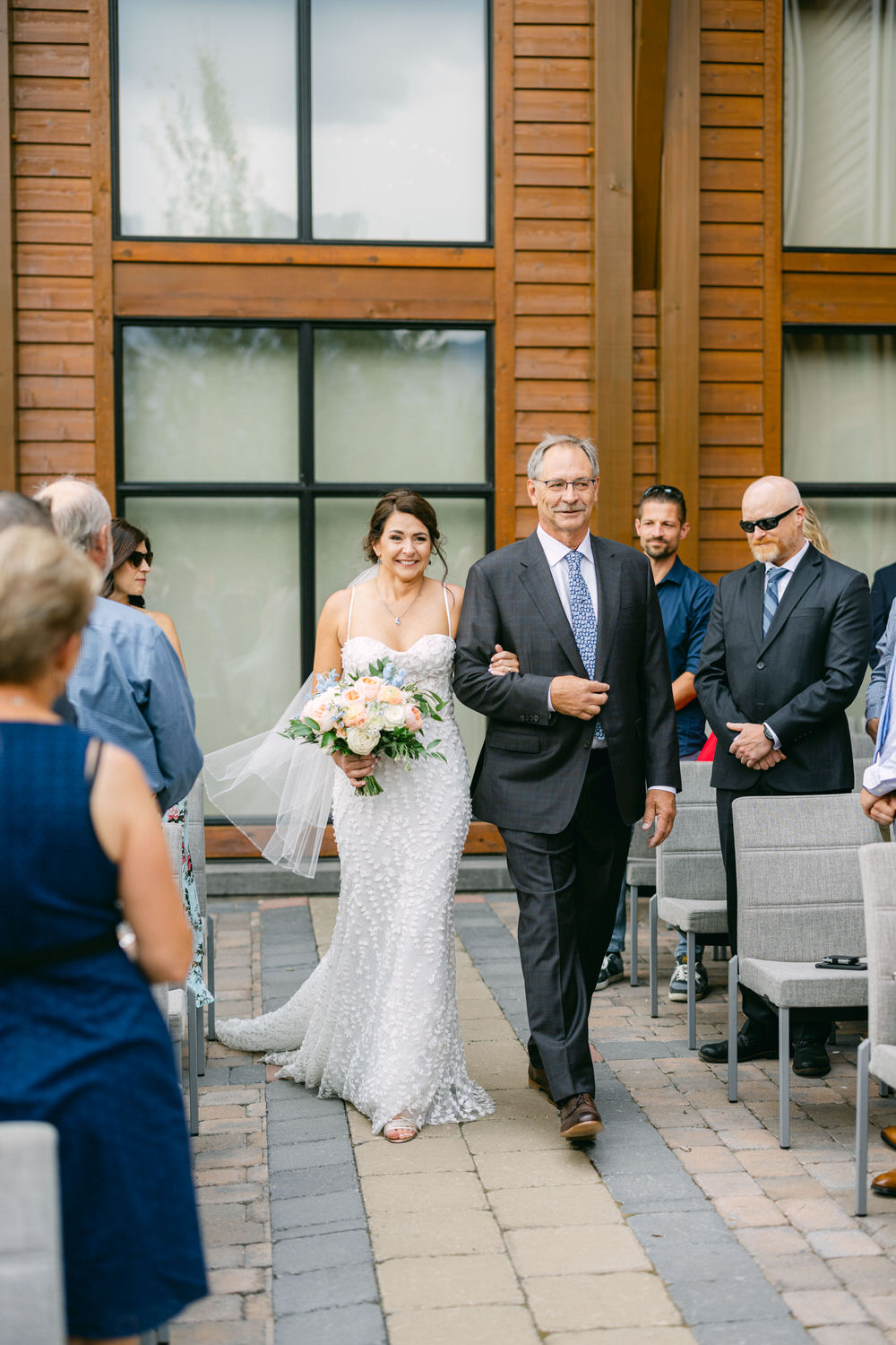 A smiling bride in a beaded white dress holds a bouquet while walking arm in arm with her father down the aisle, surrounded by wedding guests.