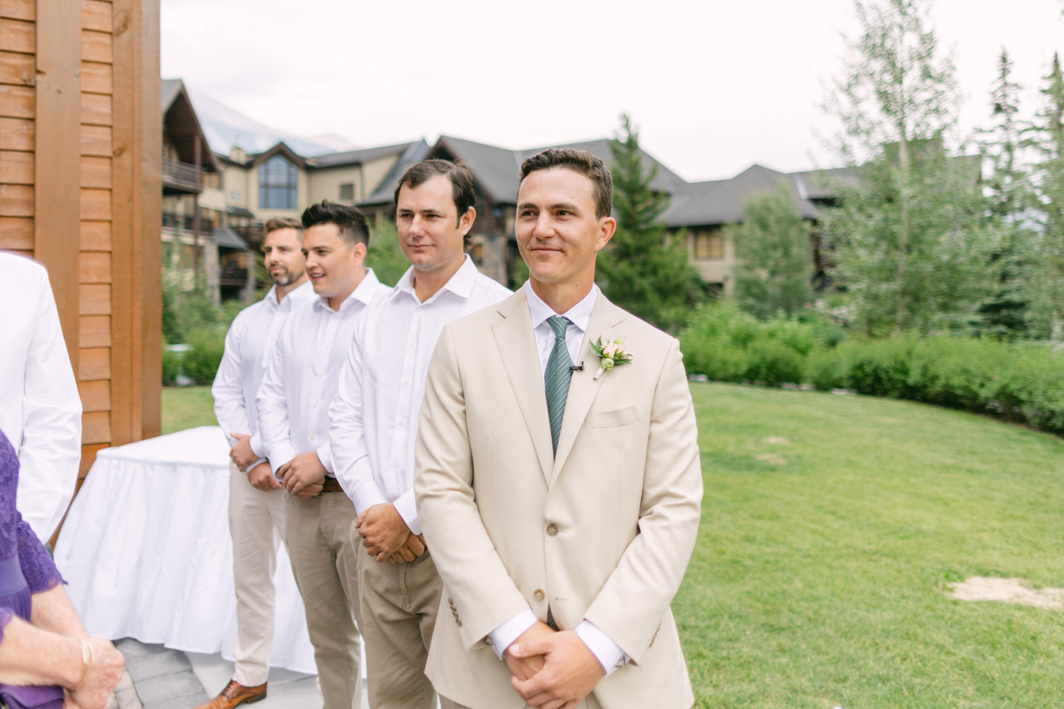 Groom and groomsmen dressed in formal attire stand outdoors at a wedding venue, with lush greenery and rustic buildings in the background.