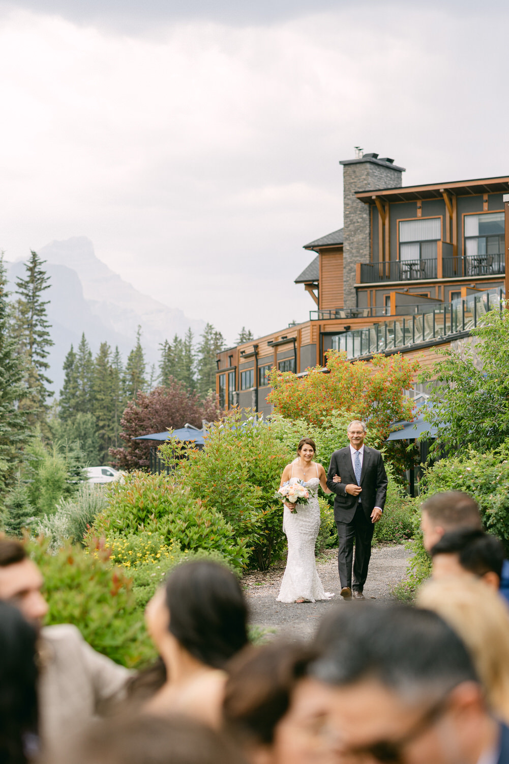 Wedding Processional Amidst Scenic Mountains::A bride walks down a garden path, holding a bouquet, accompanied by her father, with a beautiful mountain backdrop and guests in attendance.