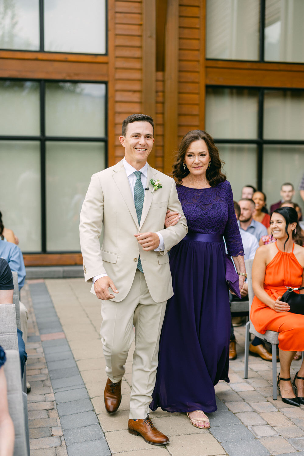 A groom smiling while walking down the aisle with his mother, surrounded by seated guests.