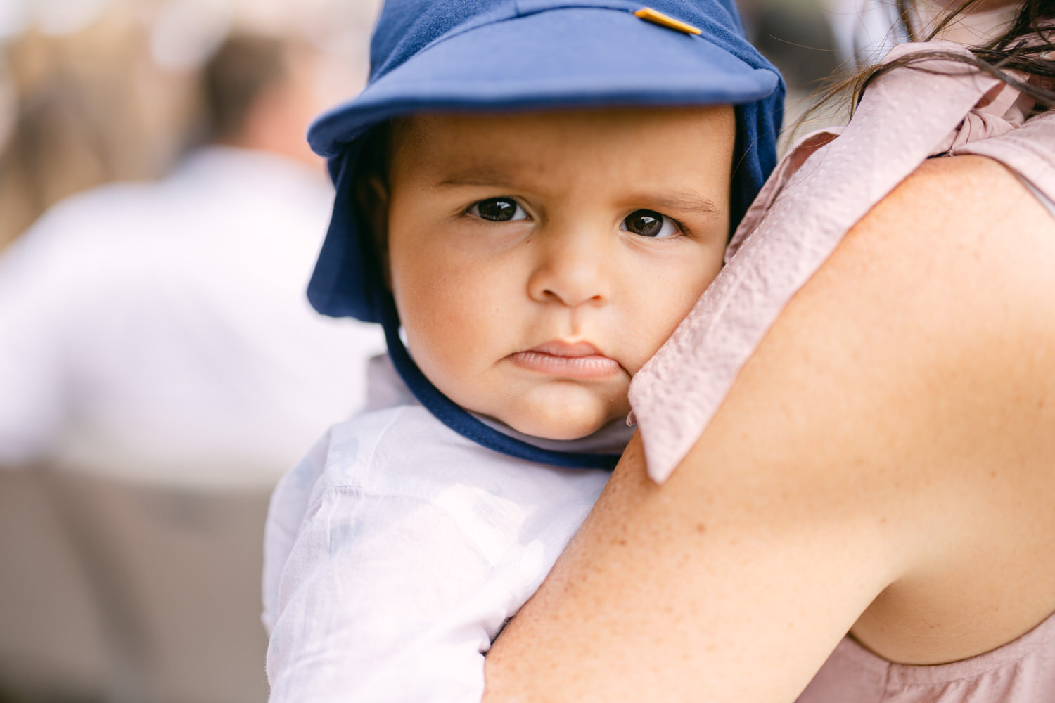 A young child with a serious expression, wearing a blue sun hat, rests against the shoulder of an adult, highlighting their close bond.