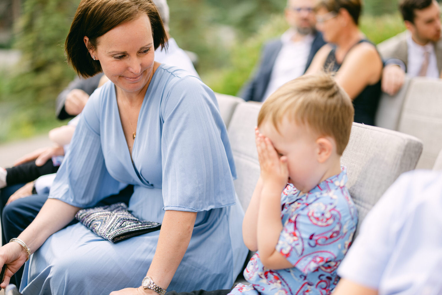 A mother smiles at her son, who covers his face with his hands, seated among guests in a casual outdoor setting.