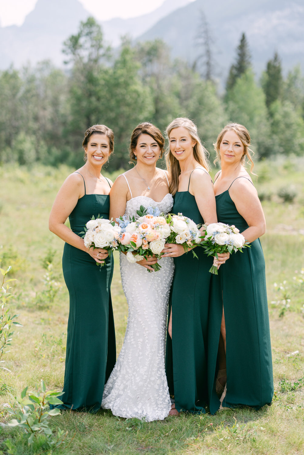 Bridal Party in Nature::Four women in elegant green dresses stand together in a scenic outdoor setting, holding beautiful flower bouquets, with mountains in the background.