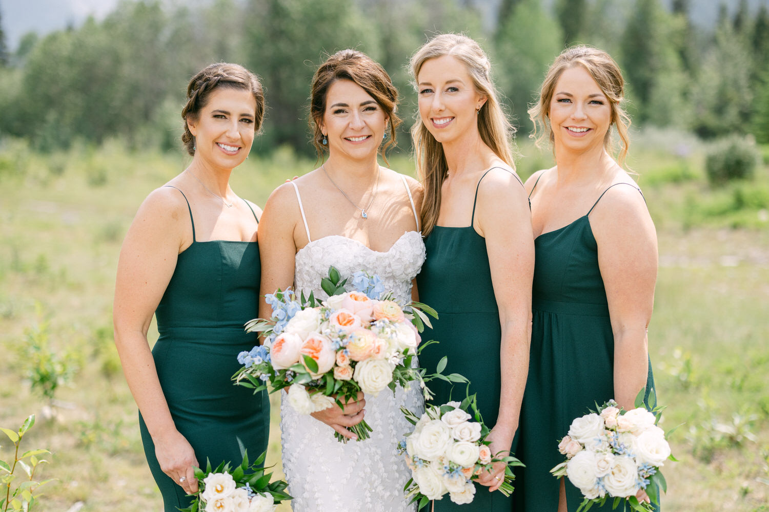 Bridal Party in Nature::Four women, including the bride in a white dress, pose together in a scenic outdoor setting, holding beautiful bouquets.