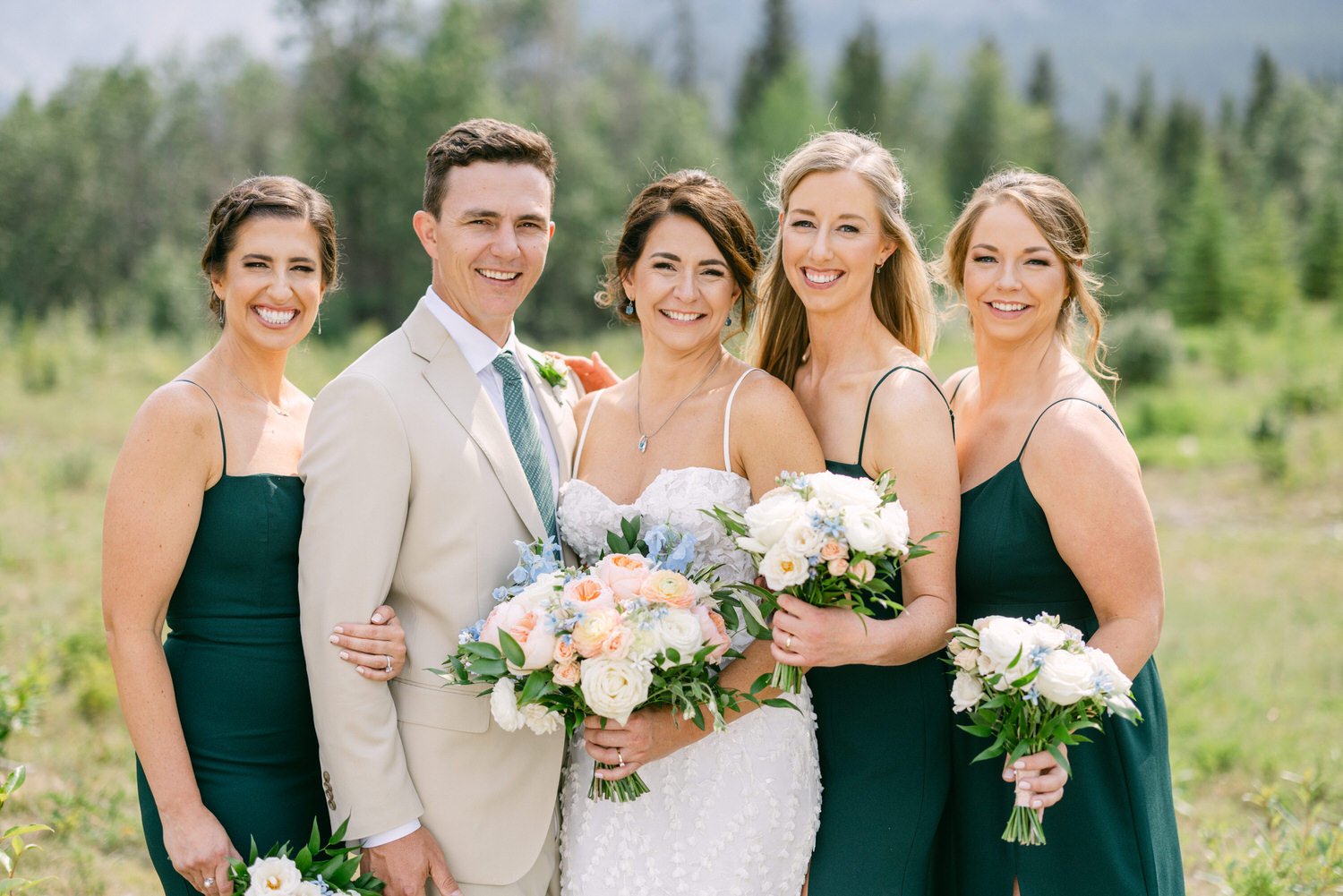 A joyful wedding party featuring the bride and her bridesmaids, all smiling and holding bouquets, set against a scenic outdoor backdrop with greenery.