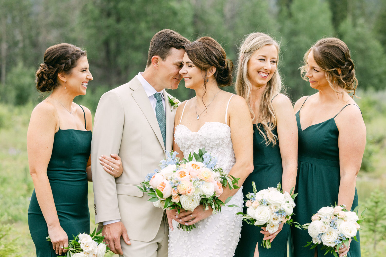 A joyful wedding moment featuring a bride and groom surrounded by their bridesmaids, all smiling amidst a lush green backdrop. The bride is holding a beautiful floral bouquet, dressed in an elegant wedding gown, while the bridesmaids are in coordinating green dresses.