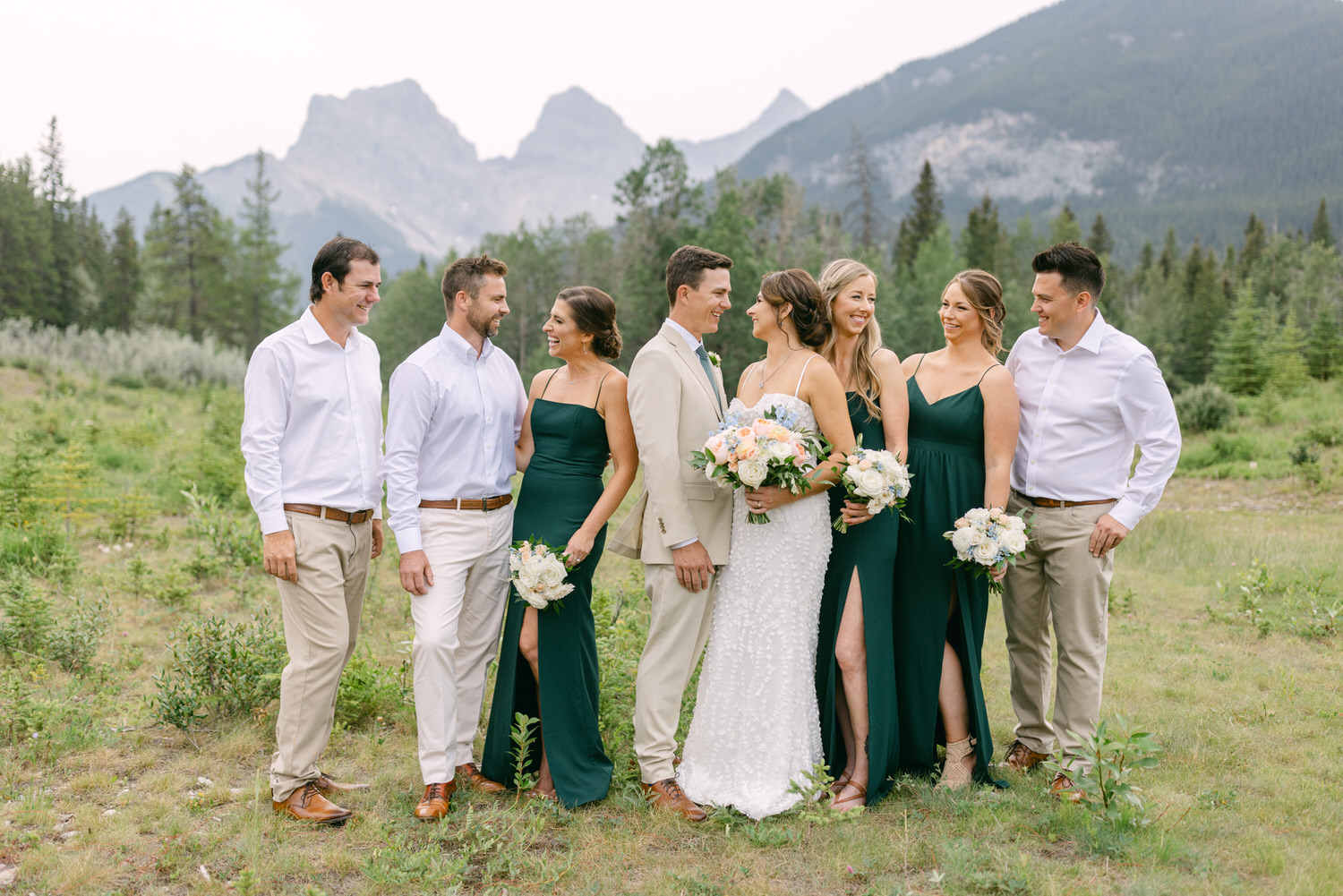 A joyful wedding party in elegant attire smiles together outdoors, surrounded by lush greenery and majestic mountains.