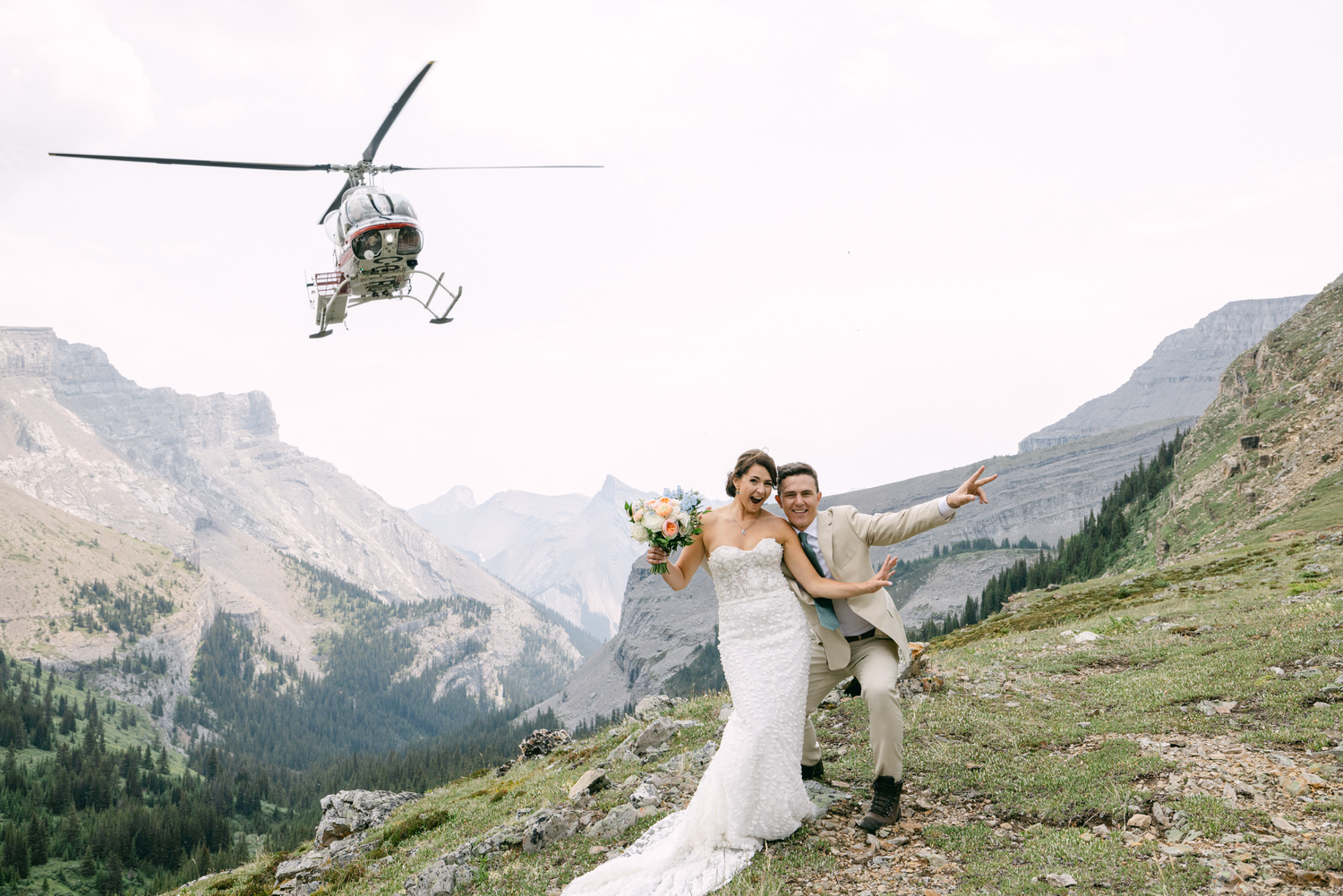 A joyful couple poses on a mountain slope, with a helicopter flying above them, surrounded by stunning mountainous scenery.