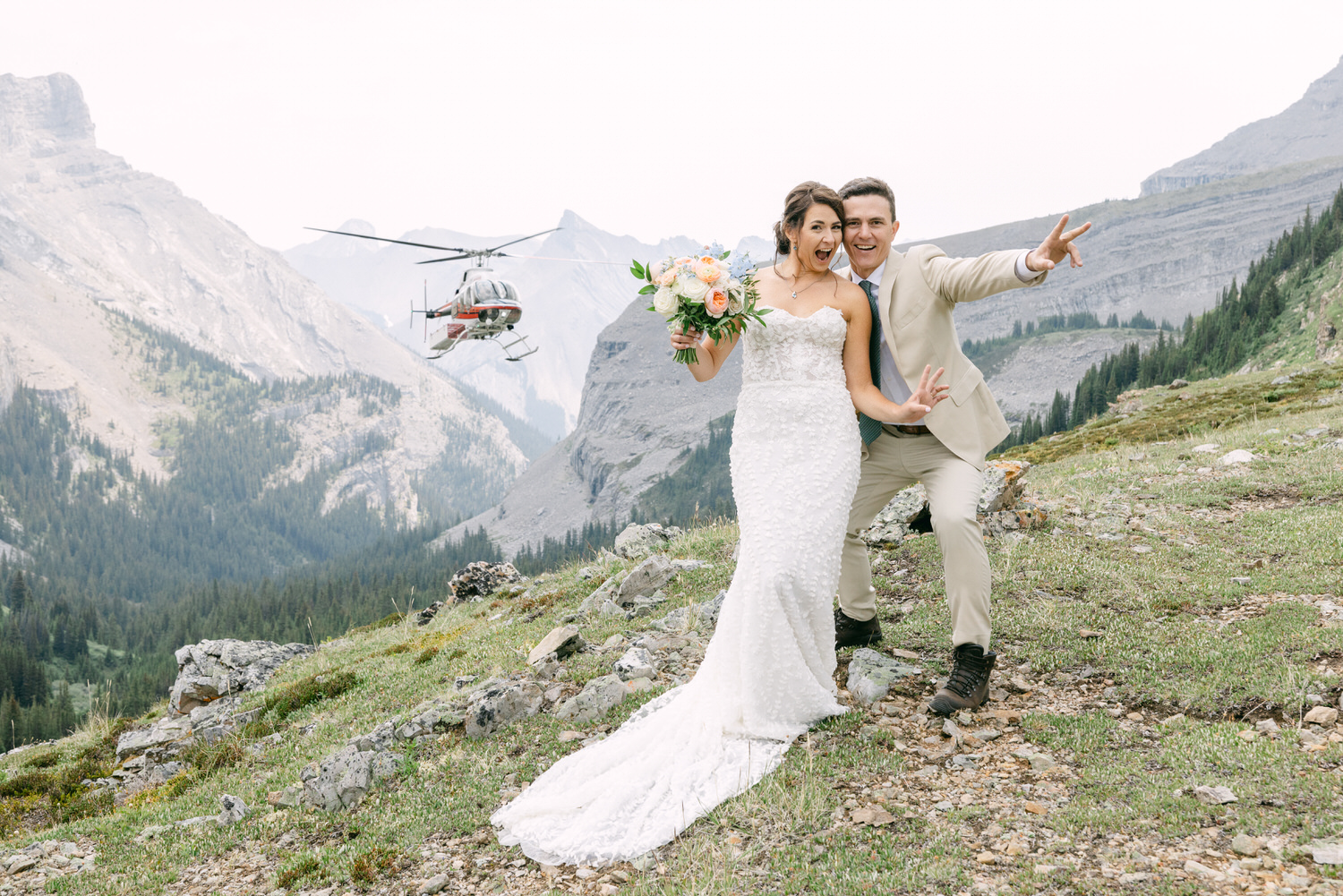 A joyful couple poses with a bouquet and playful expressions, as a helicopter hovers in the scenic backdrop of rugged mountains.