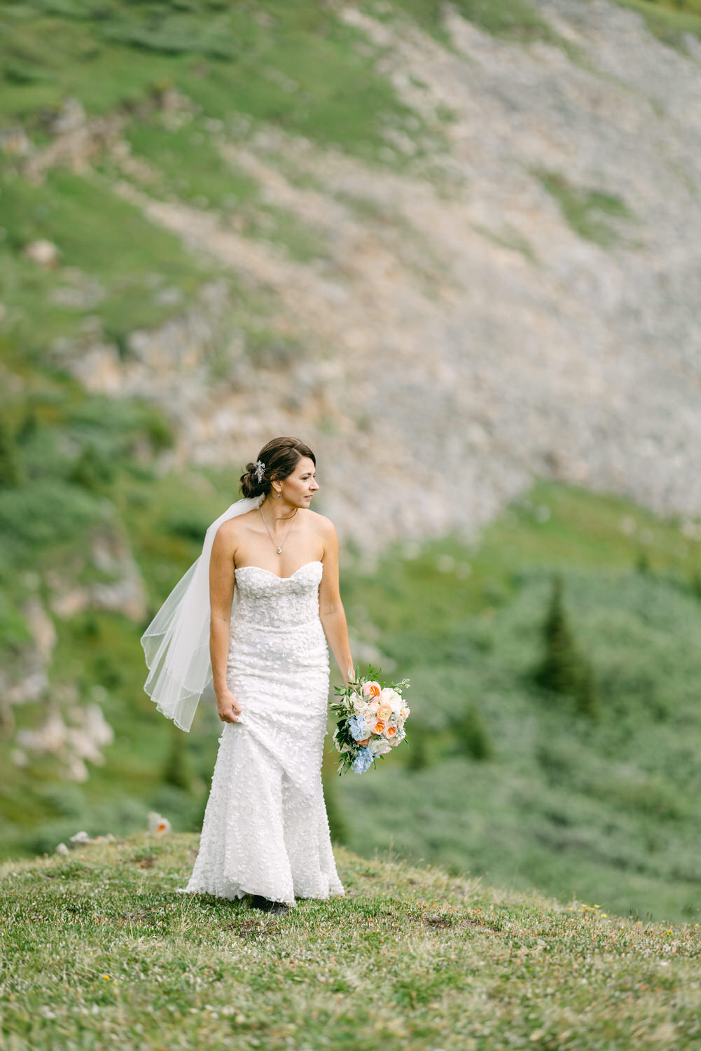 A bride in a delicate white gown stands gracefully on a lush hillside, holding a colorful bouquet, with a serene landscape in the background.