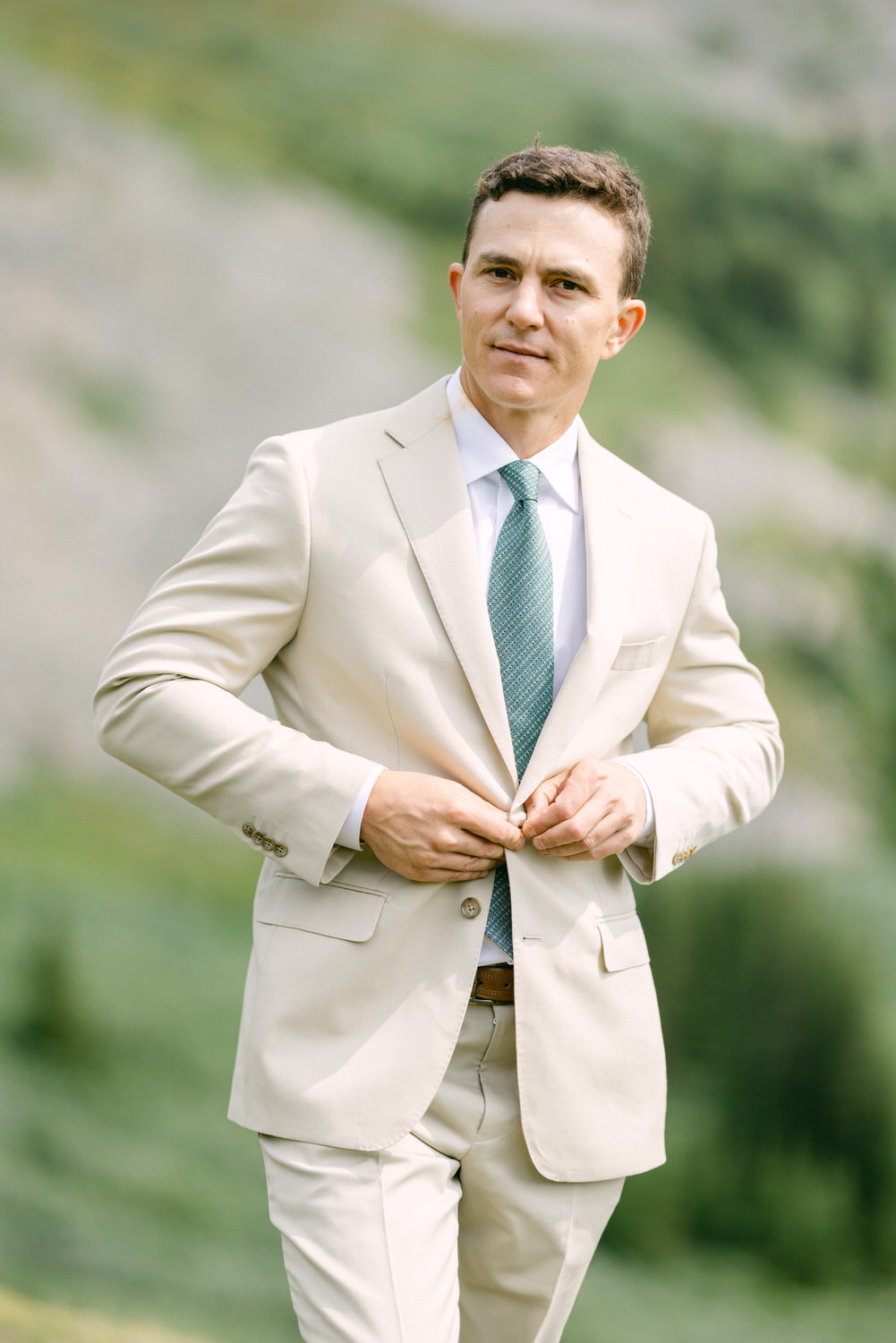 A man adjusts his suit jacket in an outdoor setting, wearing a light beige suit and a green tie, with a blurred natural background.