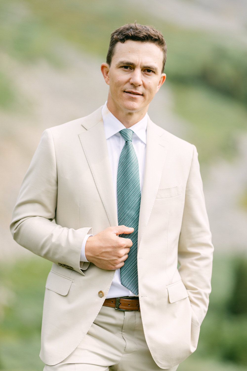 A well-dressed man in a cream-colored suit and patterned green tie stands confidently outdoors, showcasing a relaxed yet polished look.