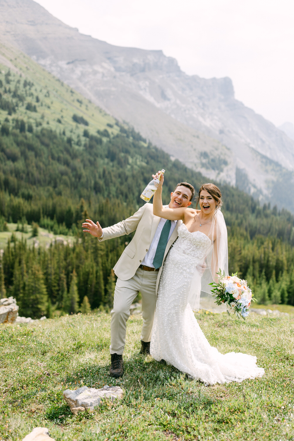 A happy couple in wedding attire celebrates outdoors, surrounded by lush greenery and mountains, with one holding a bottle and the other a bouquet.