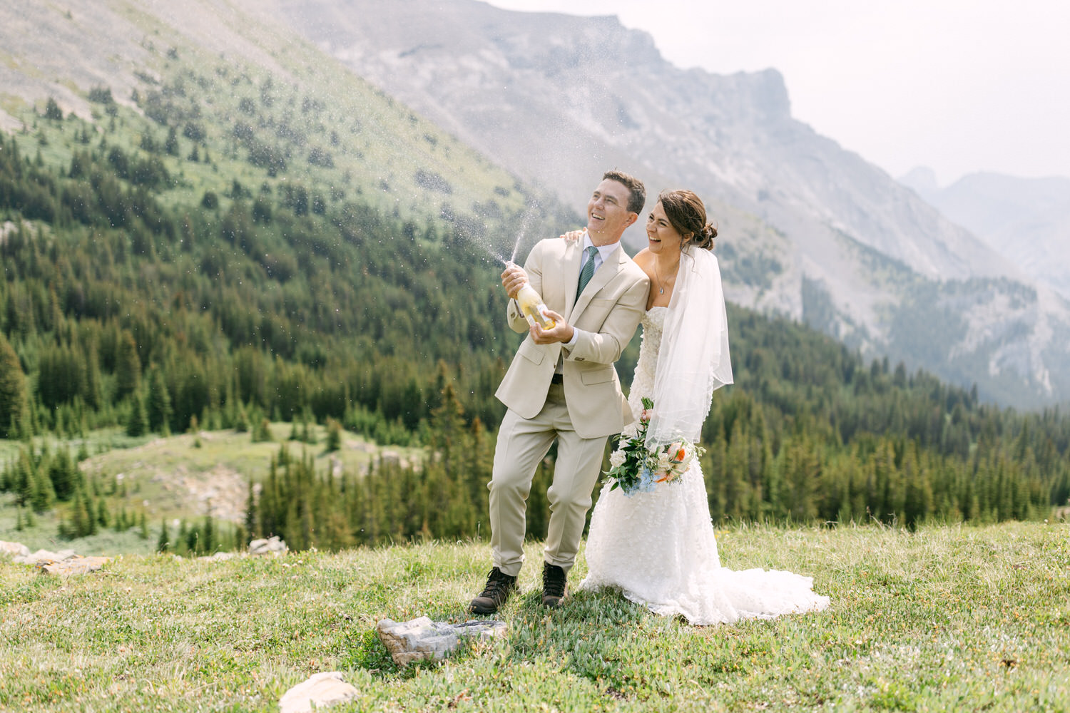 A joyful couple in wedding attire pops champagne against a stunning mountainous backdrop, capturing a moment of celebration and love.