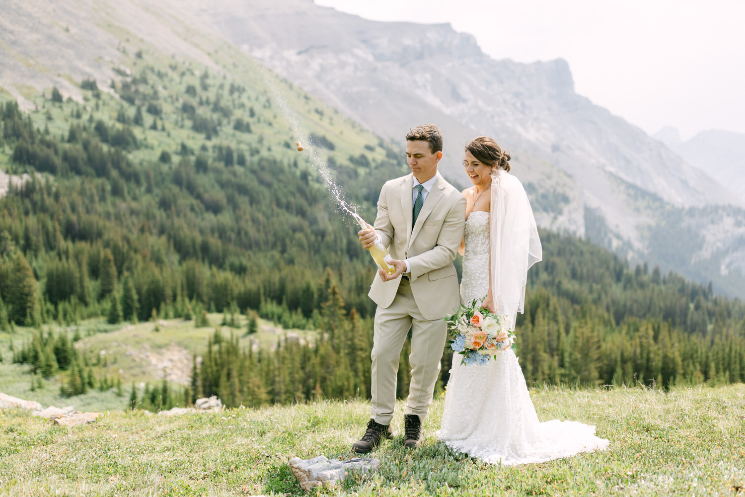 A groom popping a champagne bottle while his bride smiles, surrounded by lush green mountains and a clear sky.