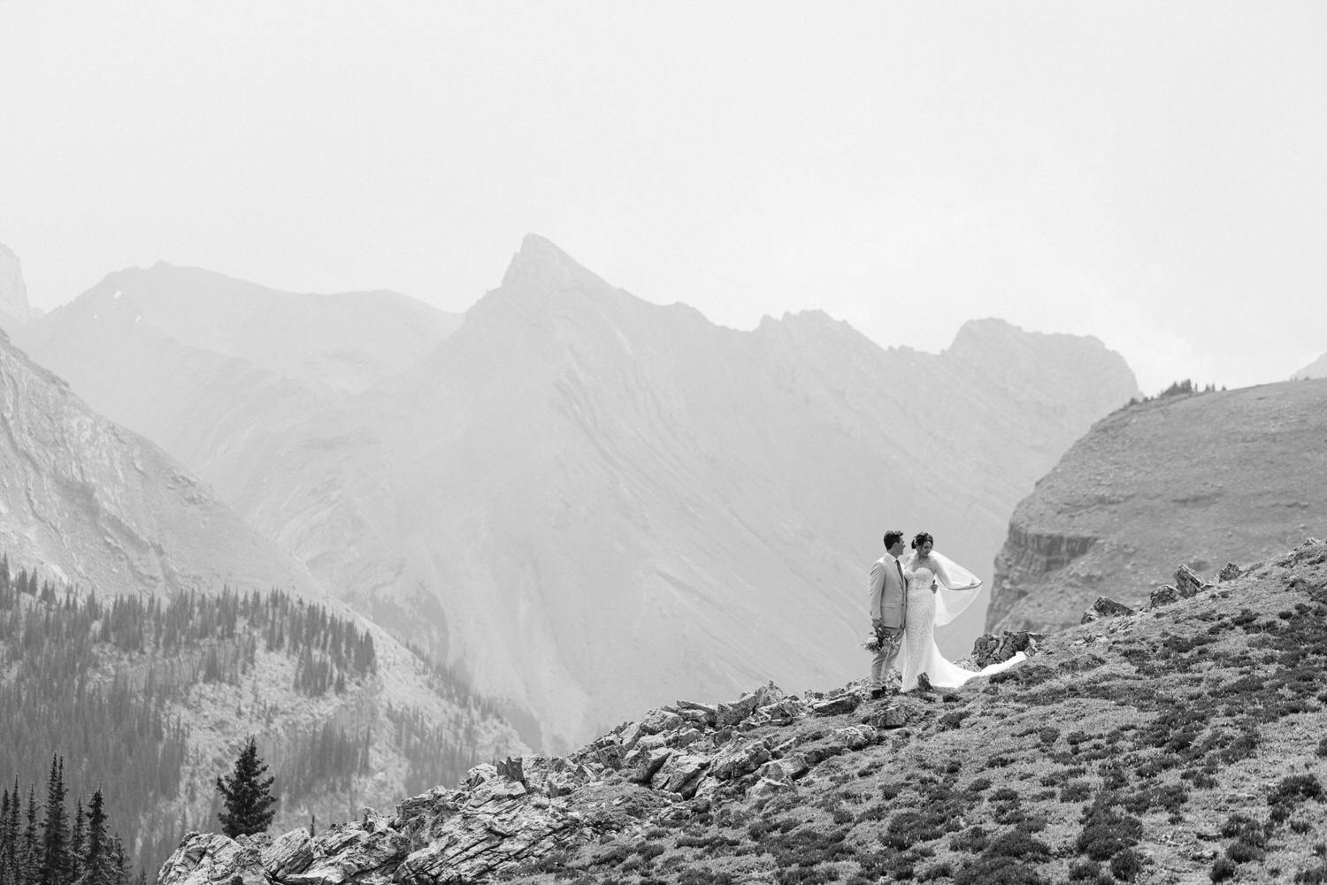 A couple stands together on a rocky mountain landscape, enveloped by misty peaks and greenery, captured in elegant black and white.