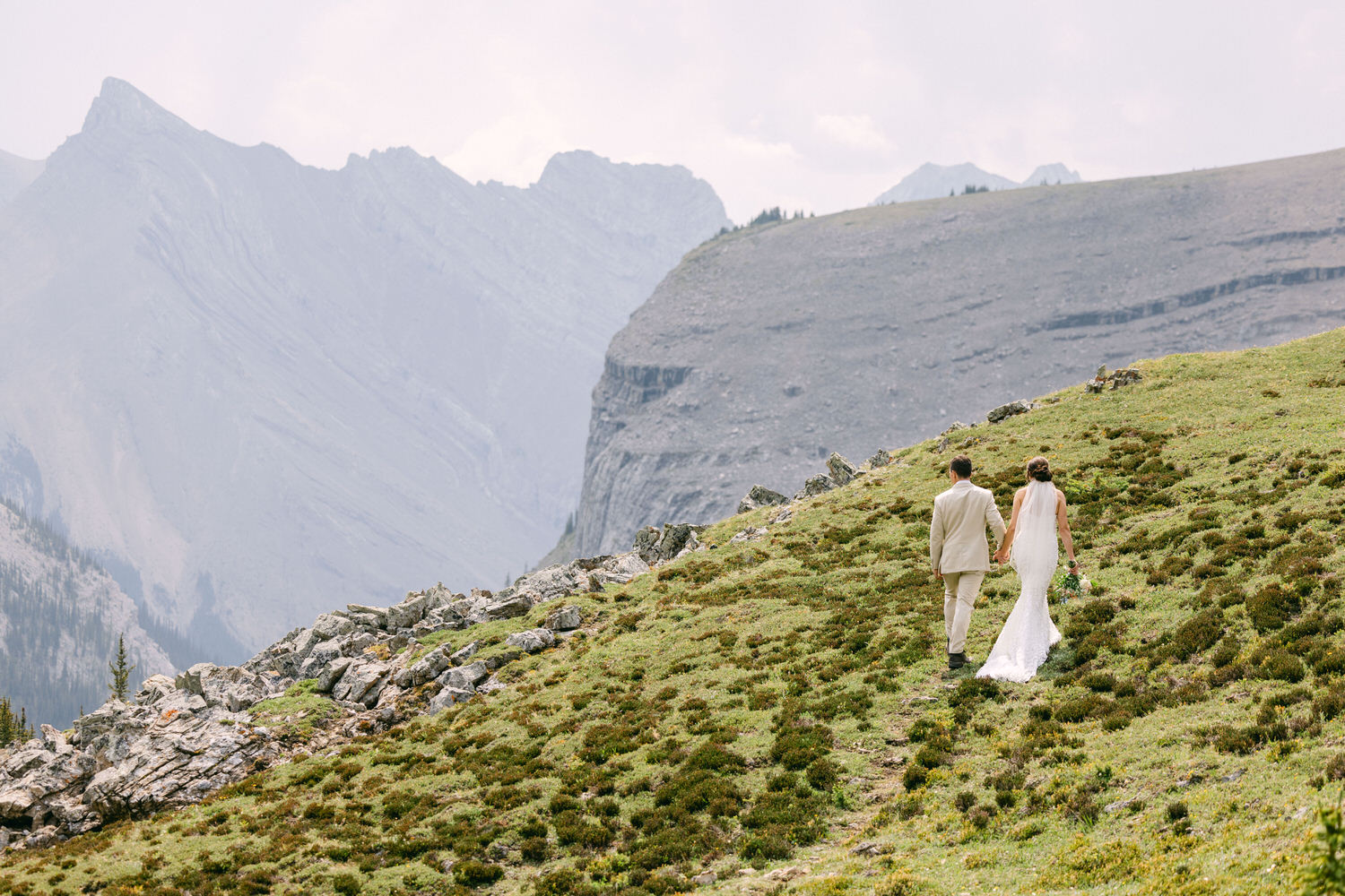 A couple in wedding attire walking hand in hand on a grassy hillside with mountains in the background.