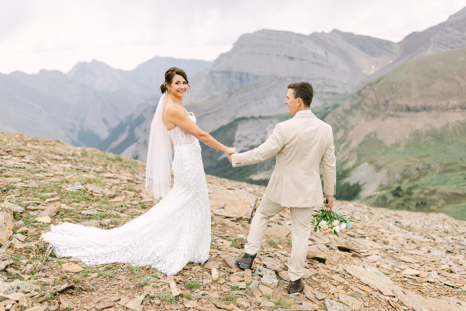 A bride and groom hold hands on a rocky mountain landscape, surrounded by stunning peaks and a serene ambiance.