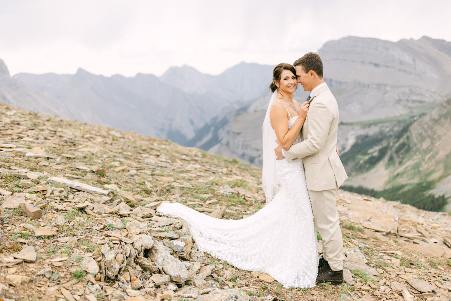 A couple embracing on a rocky mountain landscape, with mountains in the background and the bride in a beautiful white dress.