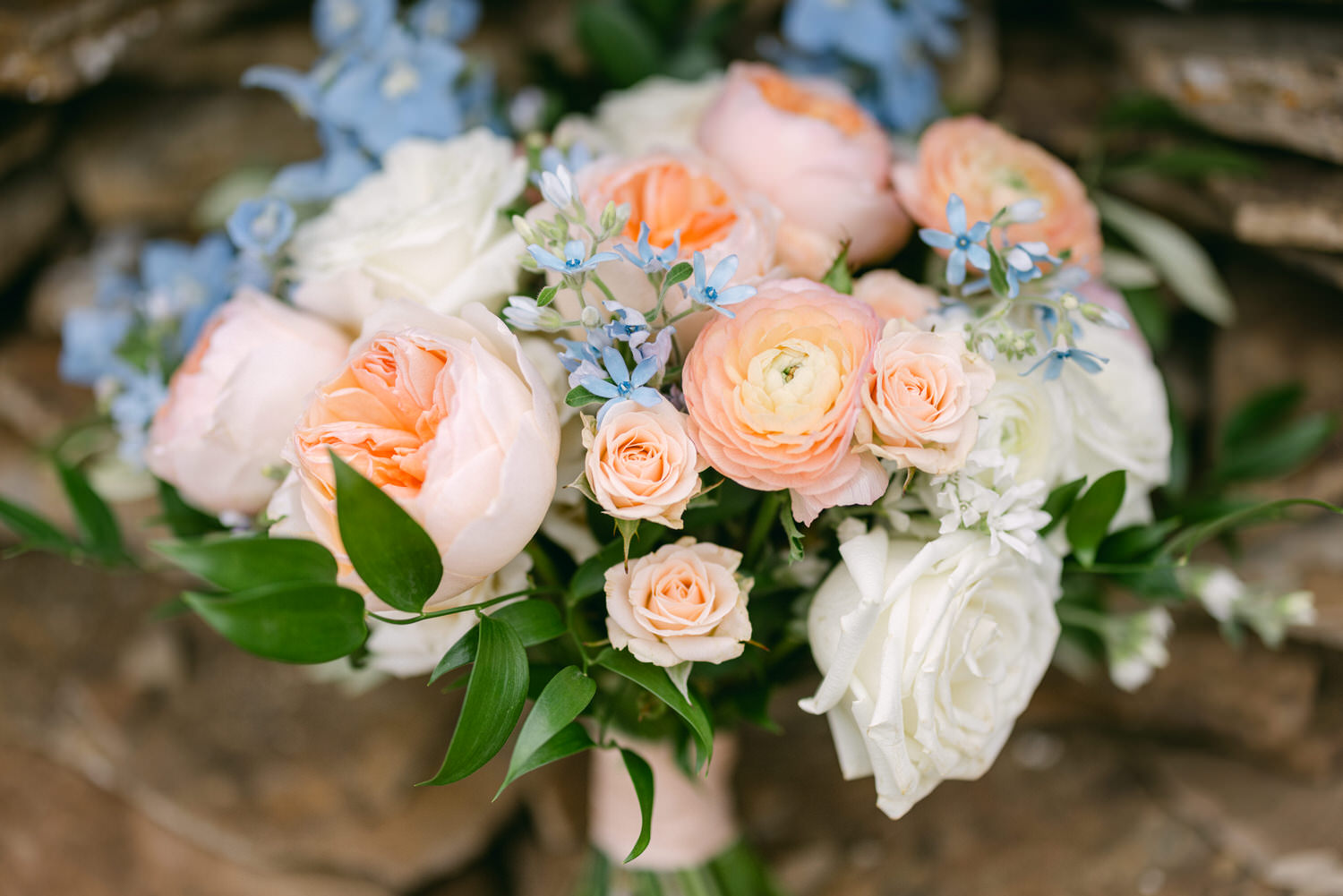 A beautiful bouquet featuring soft pink roses, delicate blue flowers, and lush greenery, arranged against a rustic stone backdrop.