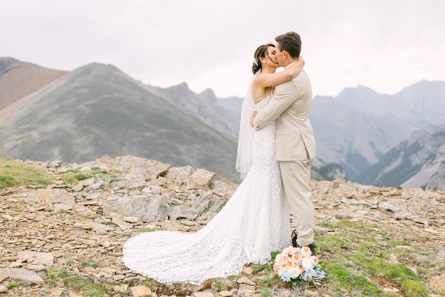 A bride and groom share a tender kiss amidst a stunning mountain landscape, surrounded by rocky terrain and majestic peaks.