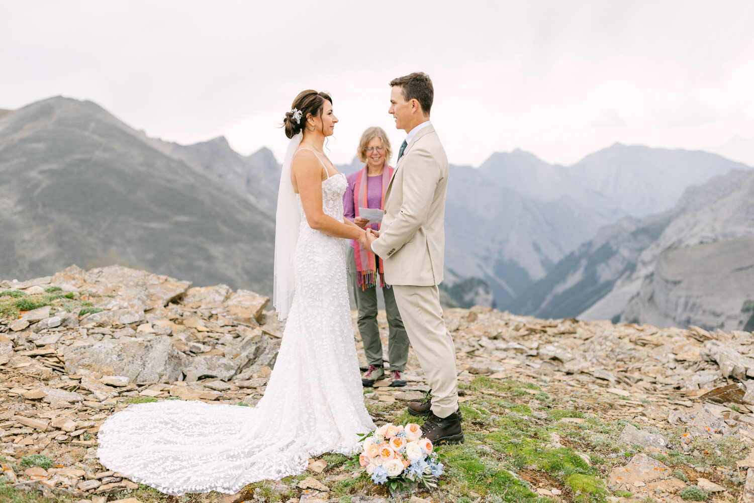 A couple exchanges vows during an outdoor wedding ceremony on a rocky mountain slope, with a scenic backdrop of distant peaks and a guest observing.