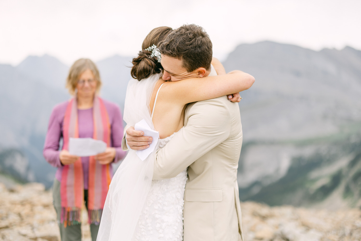 A bride and groom share an emotional embrace during their wedding ceremony in a mountainous setting, with an officiant in the background holding a script.