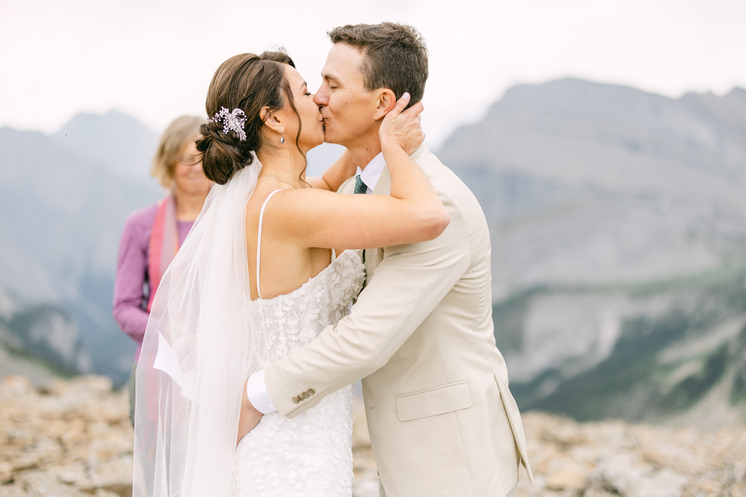 A bride and groom share a romantic kiss during their wedding ceremony in the mountains, with a serene landscape in the background.