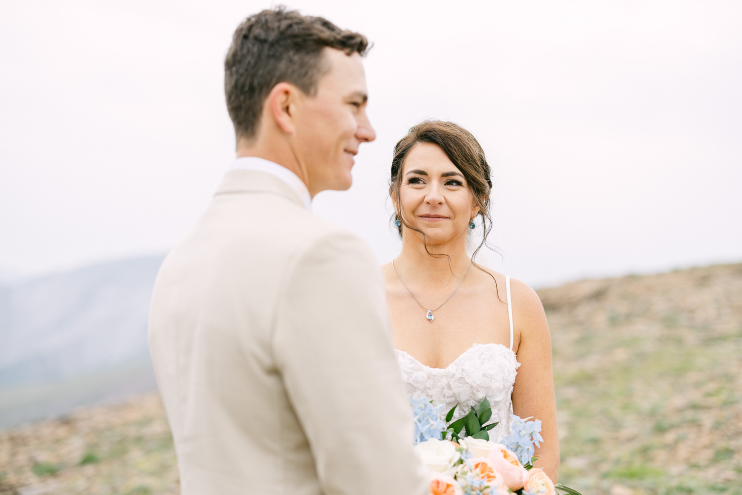 A bride in a floral dress gazes lovingly at the groom in a beige suit during an outdoor wedding ceremony amidst a scenic landscape.