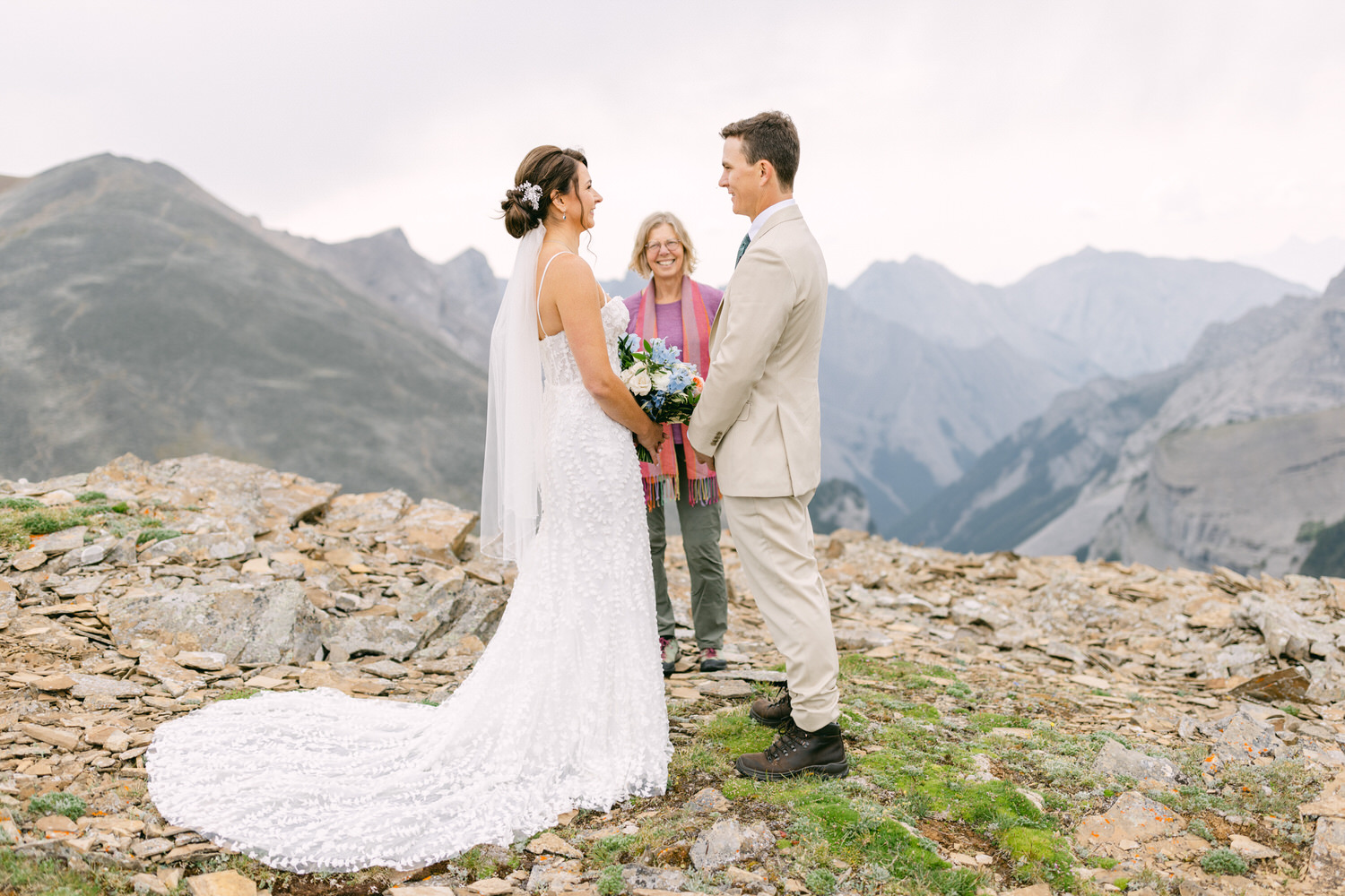 A bride in a white dress and a groom in a beige suit exchange vows on a mountain peak, with a scenic backdrop of rocky hills and a celebrant standing between them.