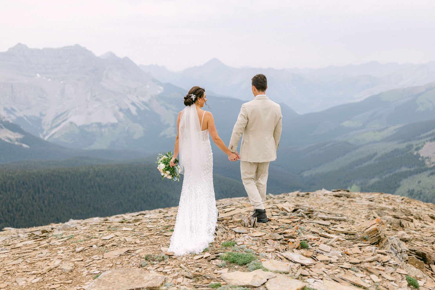 A couple holding hands and walking together on a rocky mountain summit, surrounded by stunning landscapes and mountains in the background.