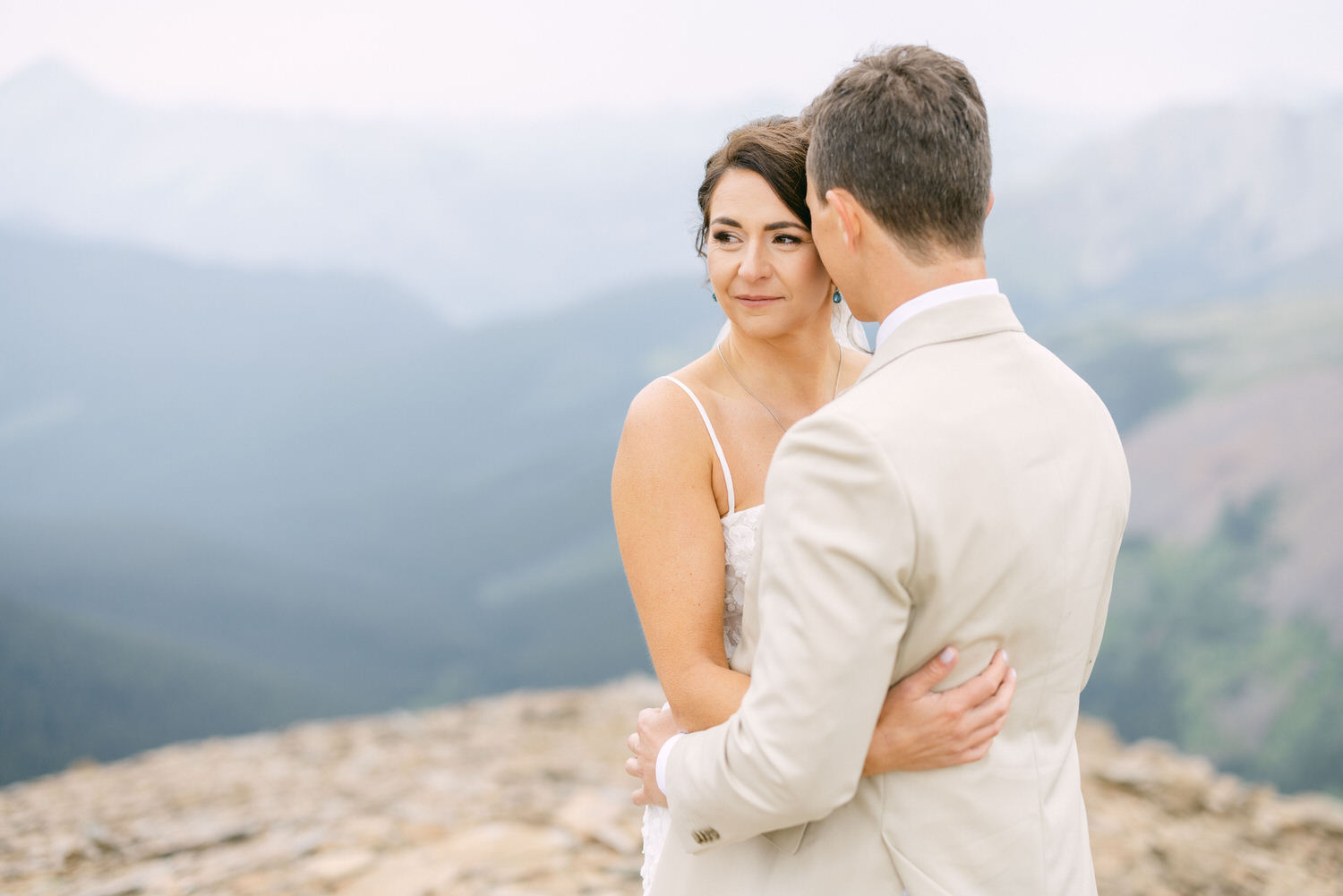 A bride and groom embrace on a rocky ledge, surrounded by lush mountains and a misty backdrop, capturing a romantic moment in nature.