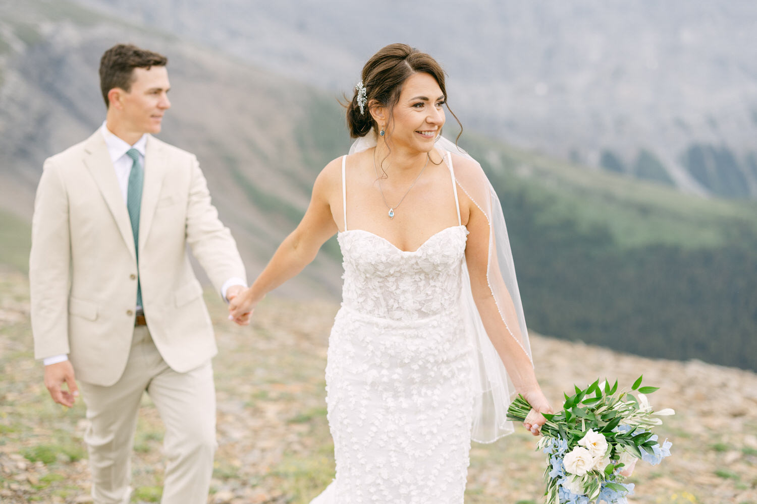 A joyful bride and groom walk hand in hand through a mountainous landscape, with the bride in a floral white gown and holding a bouquet.